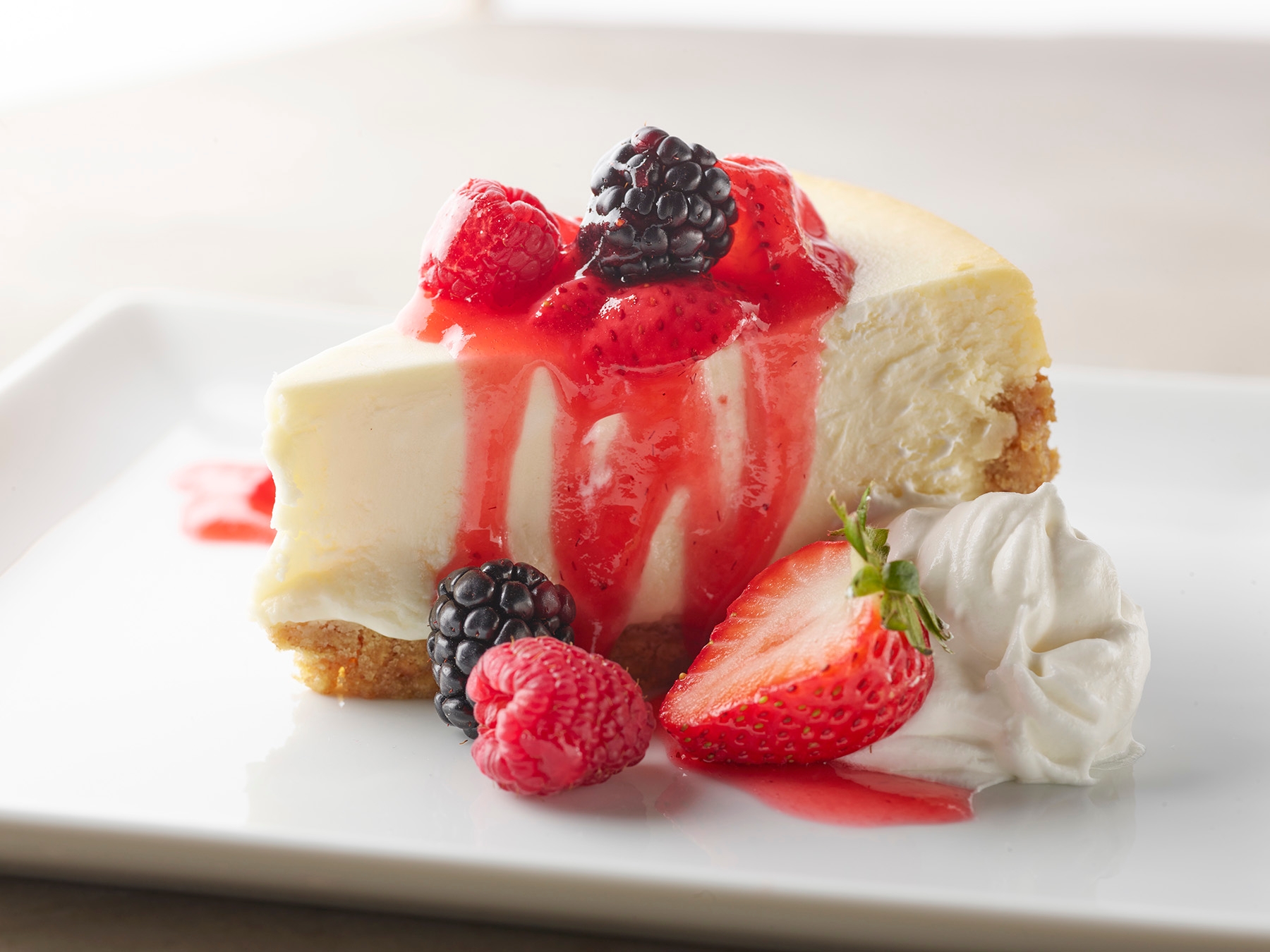 Creamy cheesecake with a graham cracker crust, topped with a decadent strawberry drizzle, mixed berries, and whipped cream.